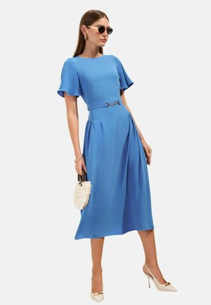 Naine sinise midi kleidiga, millel lõdvalt langevad varrukad ja vöö, hoiab valget tekstuuriga käekotti, kannab päikeseprille ja beeže kontsakingi.