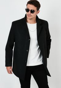 Abrigo de mezcla de lana negra con cuello estructurado, cierre de botones y bolsillos laterales, combinado con una camisa blanca y pantalones negros.