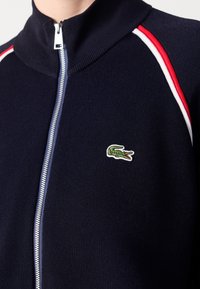 Veste zippée bleu marine avec col côtelé, bandes rouges et blanches sur les épaules, et petit logo crocodile vert sur la poitrine.
