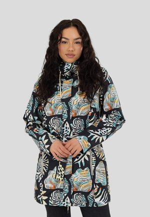 Donna con capelli lunghi e ricci che indossa una giacca multicolore con motivo animalier, mentre tiene la cerniera vicino alla vita su sfondo grigio.