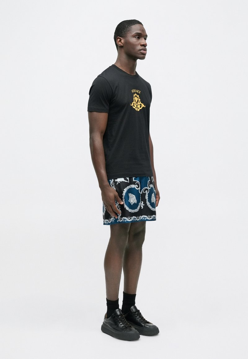 Sort bomulds-T-shirt med gyldent logo, kombineret med blå mønstrede shorts med hvide motiver og mørke sneakers med sorte såler.