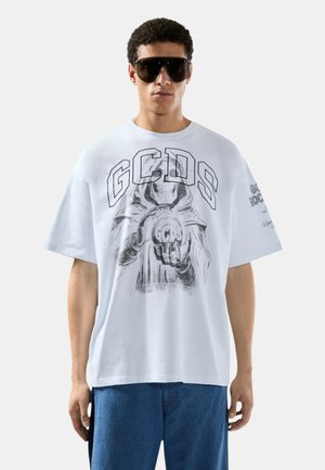 Uomo che indossa una maglietta bianca oversize con una grafica di un teschio che tiene una sfera luminosa con la scritta "GCDS", abbinata a jeans blu e occhiali da sole neri.