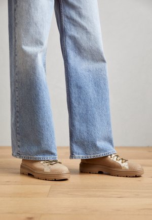 Jeans a gamba larga azzurri indossati con sneakers beige con dettagli dorati e lacci bianchi e neri su un pavimento in legno.