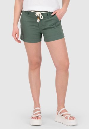 Frau trägt olivgrüne Shorts mit Seilgürtel und weiße Plateausandalen, steht vor einem schlichten Hintergrund.