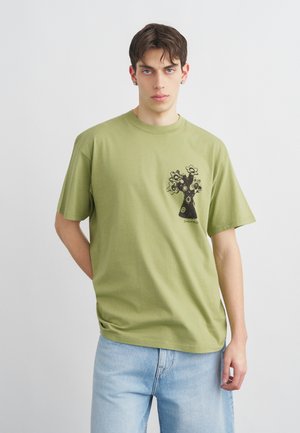 Νέος άνδρας που φοράει ανοιχτό πράσινο T-shirt με μαύρο σχέδιο ανθοφόρου δέντρου στο στήθος και ανοιχτό μπλε τζιν, στέκεται μπροστά σε απλό λευκό φόντο.