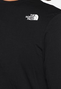 Černé tričko s dlouhým rukávem vyrobené z bavlny, s bílým logem "The North Face" na levém rameni. Hladký povrch, kulatý výstřih.
