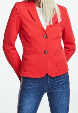 Femme portant un blazer rouge cintré avec deux poches frontales et des boutons noirs, assorti à un jean bleu à bandes latérales.