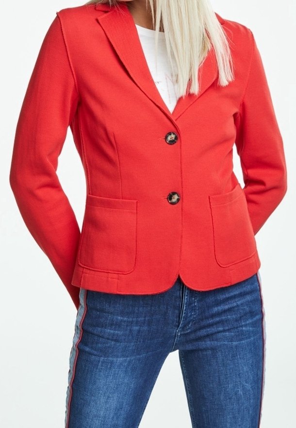 Femme portant un blazer rouge cintré avec deux poches frontales et des boutons noirs, assorti à un jean bleu à bandes latérales.