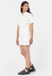 Hvit kort jumpsuit med korte ermer, glidelås foran og elastisk midje. Brukt med svarte sandaler med stropper og en tykk såle.