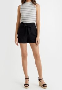 Shorts neri a vita alta con dettaglio a fiocco, abbinati a un top senza maniche grigio chiaro con righe orizzontali. Tacchi alti neri.