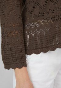 Brązowy sweter z dzianiny, z misternymi wzorami w diamenty i fale, z falbaniastym dołem i teksturowanymi rękawami, zestawiony z białymi spodniami.
