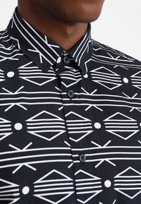 Zwarte shirt met een geometrisch wit patroon van strepen en vormen. Volledige knoopsluiting en een puntige kraag, gemaakt van lichtgewicht stof.
