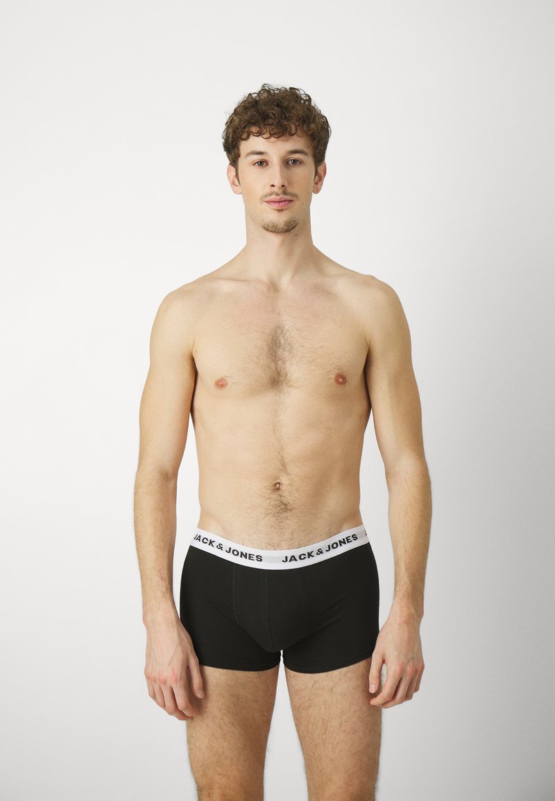 Schwarze Boxer-Briefs mit einem weißen Bund, auf dem der Text "JACK & JONES" steht. Der Stoff wirkt glatt und schmiegt sich an den Körper. Ein Mann steht mit nacktem Oberkörper.
