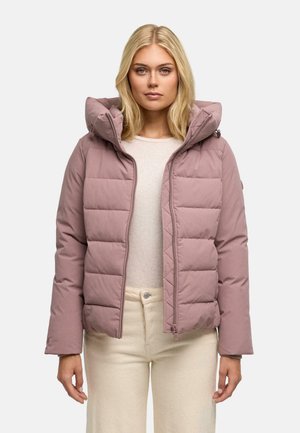 Jonge vrouw draagt een lichtpaarse gewatteerde jas met capuchon over een beige top en crèmekleurige broek, staand tegen een effen achtergrond.
