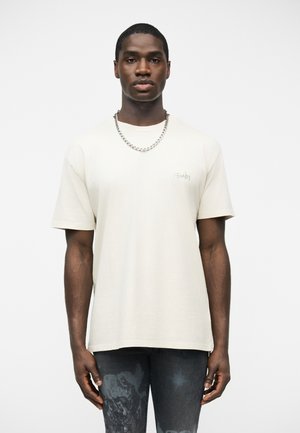 Jeune homme portant un t-shirt beige clair uni à manches courtes et une chaîne argentée, debout devant un fond blanc.