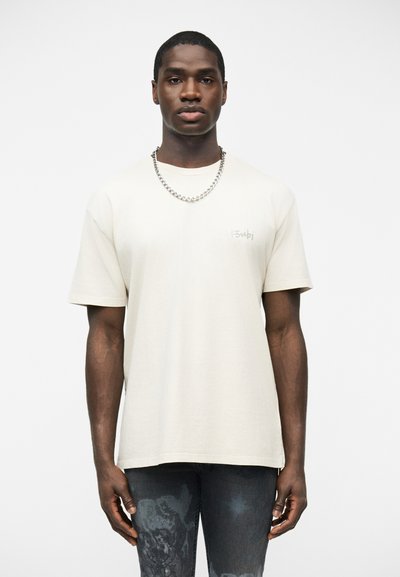 Jeune homme portant un t-shirt beige clair uni à manches courtes et une chaîne argentée, debout devant un fond blanc.