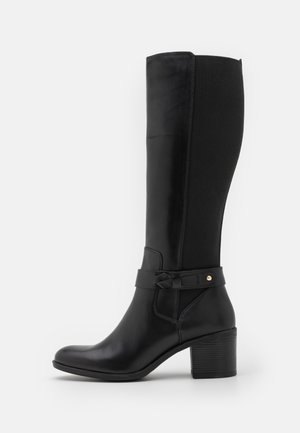 Černá kožená kotníková bota s blokovým podpatkem, elastickým zadním panelem a dekorativním páskem se zlatými cvočky na bílém pozadí.