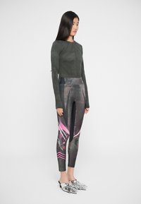 Mørkegrønn langermet topp med teksturert mønster, kombinert med svarte leggings med rosa grafiske detaljer og en blank sølvskofe.
