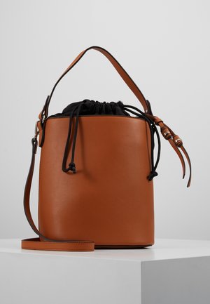 Brązowa skórzana torba typu bucket z czarnym ściągaczem, regulowanym paskiem na ramię i pojedynczym uchwytem, leżąca na białej powierzchni.