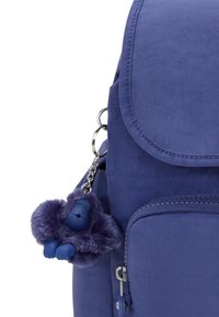 Lila tygryggsäck med klaffstängning, sidoficka och en fluffig lila gorillanyckelring fäst. Slät yta och silverfärgad hårdvara.