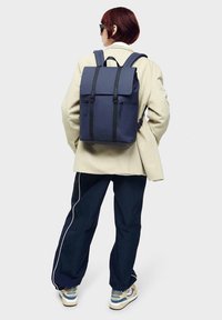 Navy backpack with a matte finish, featuring adjustable straps, flap top, black hardware, and two front zippers, worn over a light beige coat.