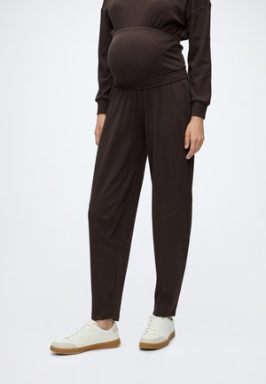 Personne enceinte portant un pantalon de maternité marron foncé et des baskets blanches, debout devant un fond uni.