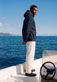 Homme en veste sombre et pantalon blanc se tenant sur un bateau, regardant une mer calme avec des falaises lointaines sous un ciel clair.
