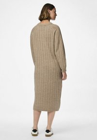 Beige stickad klänning med ribbad textur, långa ärmar och sidoklyfta. Slätt tyg sträcker sig till mitten av underbenet, ihop med avslappnade vita sneakers.