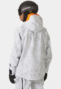 Veste de ski dans un motif camouflage gris clair, avec un col haut et une touche d'orange sur la capuche. Elle comprend des poignets ajustables et des poches zippées.