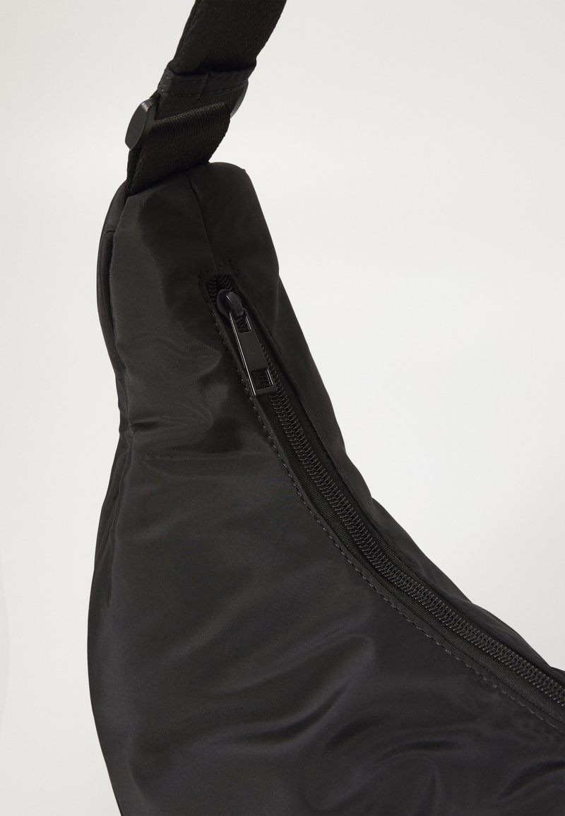 Sac à bandoulière noir en tissu lisse et mat, doté d'une poche zippée sur le côté, d'une bandoulière réglable et d'un design incurvé.