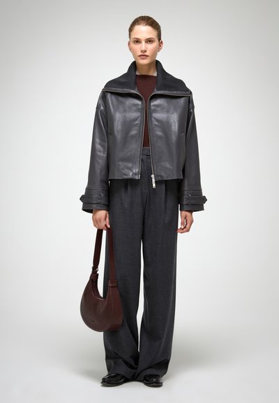 Giacca cropped in pelle nera con colletto alto, chiusura con zip, abbinata a pantaloni grigi a gamba larga e una borsa a spalla marrone.