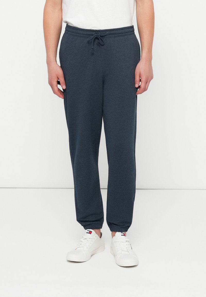 Bărbat îmbrăcat în pantaloni jogger gri închis, cu talie elastică și manșete elastice, asortați cu adidași albi și un tricou alb.