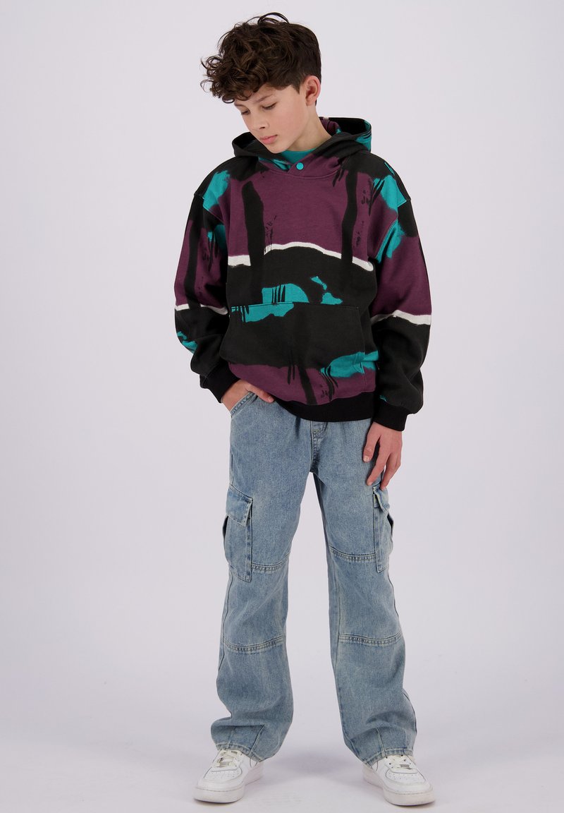 Hettegenser i lilla, svart og teal med abstrakte mønstre; paret med lyseblå cargo-jeans og hvite sneakers.
