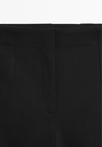 Czarne spodnie na miarę o gładkiej fakturze, z czystym pasem i subtelnym detalem szwów. Brak widocznych kieszeni.