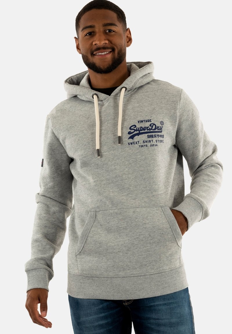 Sudadera gris de tela suave, con un bolsillo frontal, capucha con cordones y logo bordado en azul. Combinada con jeans azules.
