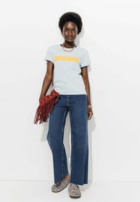 T-shirt gris avec "Horizon" en orange, jean large bleu, sac bandoulière à franges rouges et chaussures à enfiler en imprimé léopard.