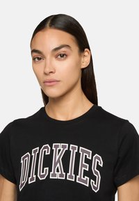 Schwarzes Baumwoll-T-Shirt mit einem auffälligen "DICKIES"-Logo in Creme- und Dunkellila-Tönen, klassischem Rundhalsausschnitt und kurzen Ärmeln.