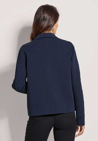 Marineblaue Jacke mit Kragen, langen Ärmeln und lockerer Passform. Glattes Material mit sichtbaren Stickdetails an den Schultern und Bündchen.