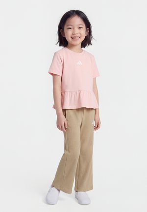 Usmievajúce sa dieťa stojace čelom vpred, oblečené v ružovej košeli s volánmi, béžových manšestrových nohaviciach a bielych slip-on topánkach na bielom pozadí.