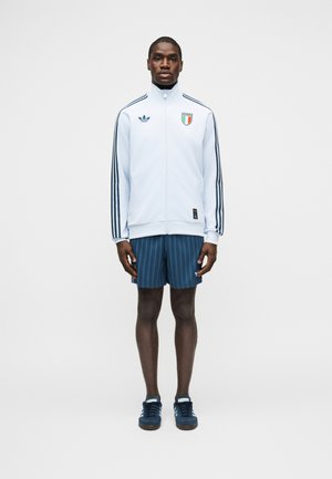 Hombre de pie mirando hacia adelante, vistiendo una chaqueta deportiva blanca de Adidas con rayas y el escudo de Italia, pantalones cortos azules a rayas y zapatillas azules.