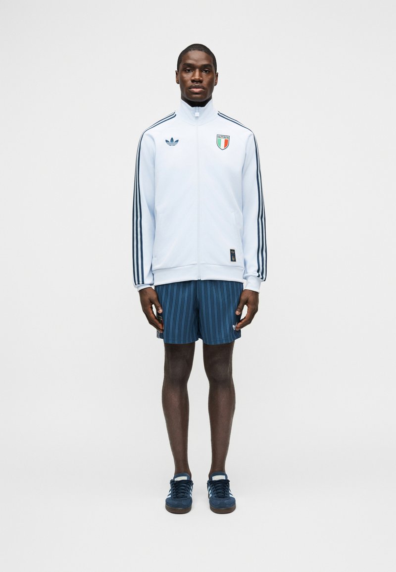 Mand stående vendt fremad iført hvid Adidas træningsjakke med striber og Italiens våbenskjold, blå stribede shorts og blå sneakers.