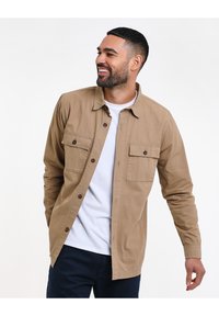 Tan button-up blouse met twee borstzakken, gecombineerd met een witte ronde hals t-shirt. Glad katoenmateriaal, ontspannen pasvorm, volledige knoopsluiting vooraan.