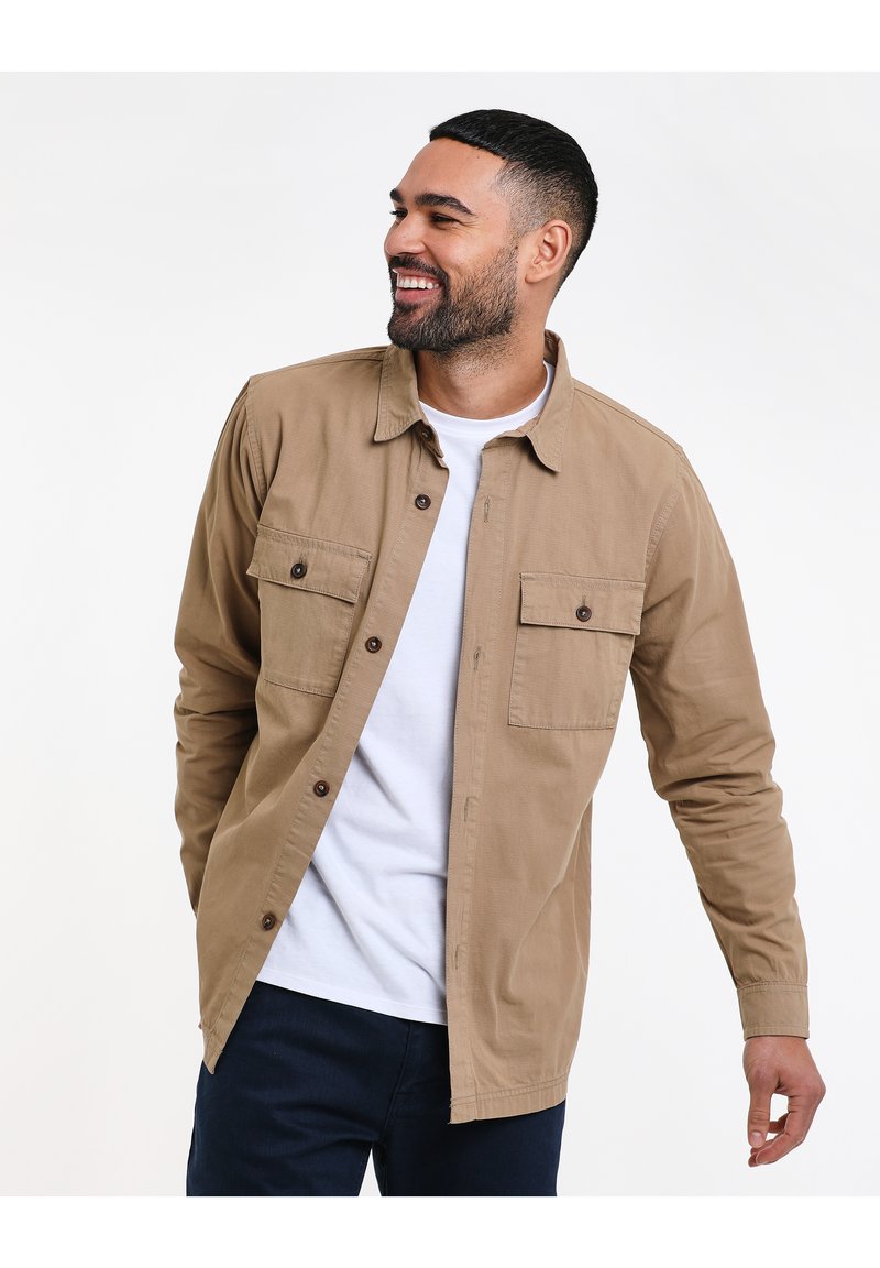 Tan button-up blouse met twee borstzakken, gecombineerd met een witte ronde hals t-shirt. Glad katoenmateriaal, ontspannen pasvorm, volledige knoopsluiting vooraan.
