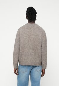 Pulover tricotat de culoare maro deschis, cu o croială lejeră, guler rotund și manșete cu model striat. Asortat cu blugi din denim de culoare albastru deschis.