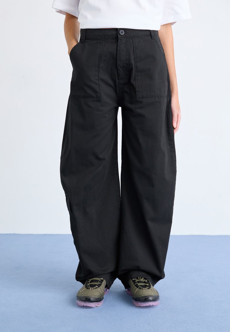 Schwarze, weit geschnittene Hosen aus Baumwolle mit vorderen Taschen, einem Knopfverschluss und sichtbaren Nähten. Kombiniert mit Sportschuhen.