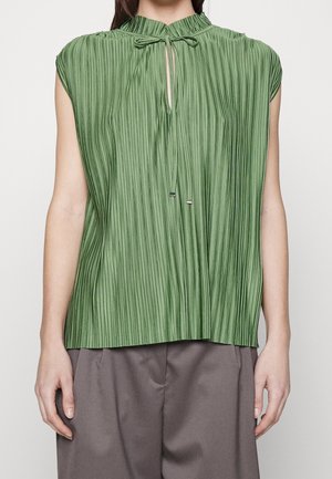 Blouse verte plissée sans manches avec un col haut froncé et un détail œillet noué, portée avec un pantalon gris ample.