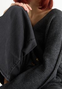 Přes oversized tmavě šedý svetr s texturovanou tkaninou a výstřihem do V, který je vrstvený přes tmavé džíny. U pasu se nachází několik knoflíků.