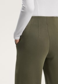 Olive zelene široke hlače z gladko strukturo, ki vključujejo štiri žepke in vertikalne gubice. V kombinaciji z oprijetim belim dolgim topom.