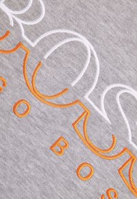 Šedá látka s texturovaným povrchem, zdobená oranžovým a bílým vyšívaným písmem v moderním písmu. Akcenty zvýrazňují název značky.