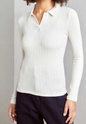 Femme portant un polo blanc ajusté à manches longues, avec trois boutons, associé à un pantalon bleu marine sur un fond neutre.